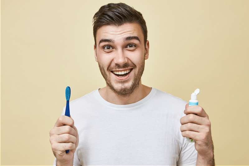 energetic-positive-young-man-with-stubble-posing-with-toothbrush-whitening-paste-smiling-broadly-with-perfect-white-teeth-healthy-habits-daily-routine-dental-care